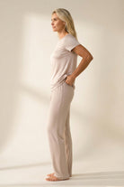 A woman stands in profile wearing a light beige short-sleeve t-shirt and matching wide-leg pajama pants against a warm off-white studio background.