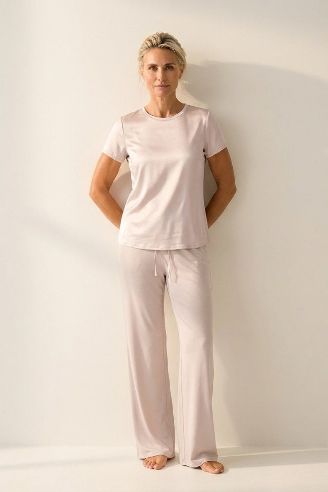 A woman modeling a light pink short-sleeve pajama top and matching wide-leg pajama pants against a plain off-white wall.
